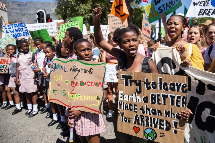 Group of people demonstrating about Climate Justice. Two young people hold placards reading: "Global warming isn't a prediction, it is happening" and "Try to leave Earth a better place than when you arrived!" respectively